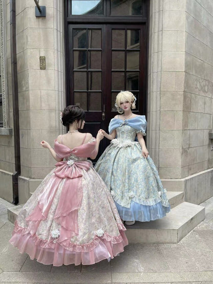 Two women wearing elaborate historical-style ball gowns posing on the steps of a stone building. The woman on the left wears a pink and white lace gown with a large bow at the back, while the woman on the right wears a light blue and white lace gown with a voluminous skirt.