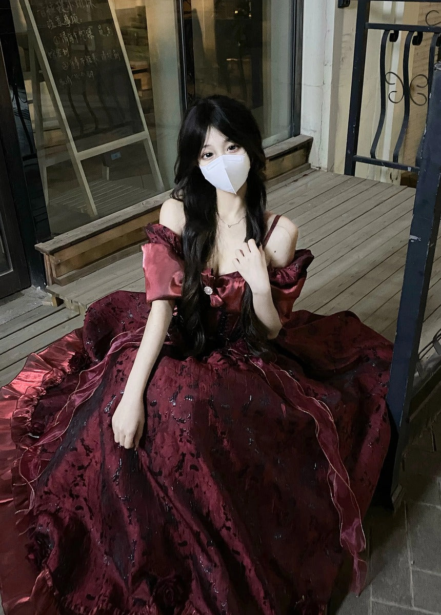 A woman wearing a voluminous dark red off-the-shoulder ball gown with a white face mask, sitting on a wooden deck in front of a building.
