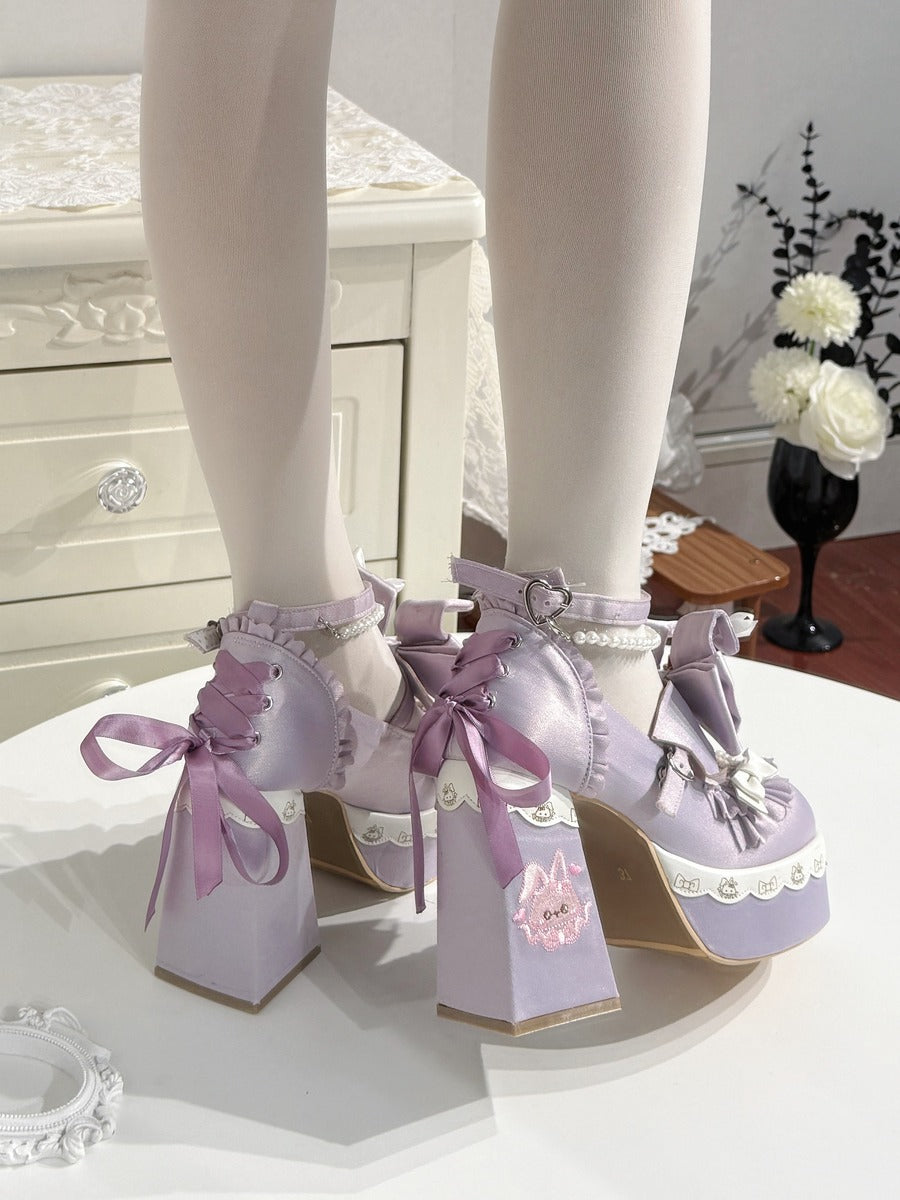 A pair of lavender platform heels with chunky block heels, lace-up ankle straps, and large bows, shown on a model's feet wearing white tights in an indoor setting.