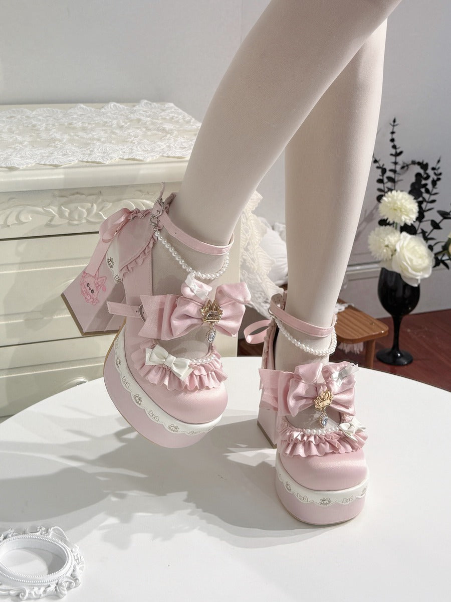 A pair of pastel pink Lolita-style platform heels with large satin bows, pearl trim, and decorative charms, shown being worn on a doll or mannequin legs in an indoor styled setting.