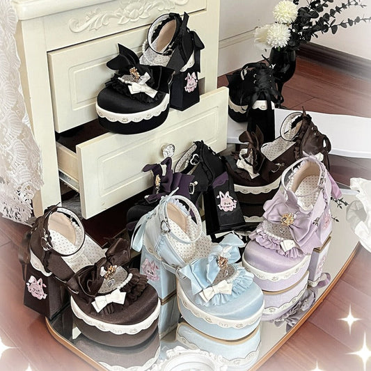 A collection of pastel Lolita-style platform shoes displayed on a mirrored tray and a small cream cabinet, featuring black, lavender, light blue, and pink designs with lace, bows, and decorative accents.