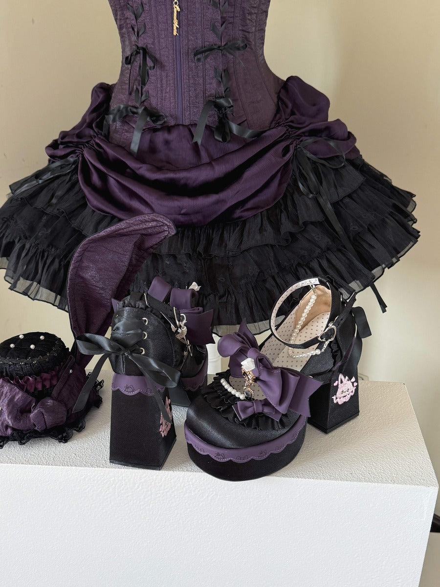 A gothic-style outfit display featuring a deep purple corset with black ruffled layers, a matching purple and black skirt, black platform shoes with purple bows, and black handbags with decorative details, arranged on a white display block against a beige wall.