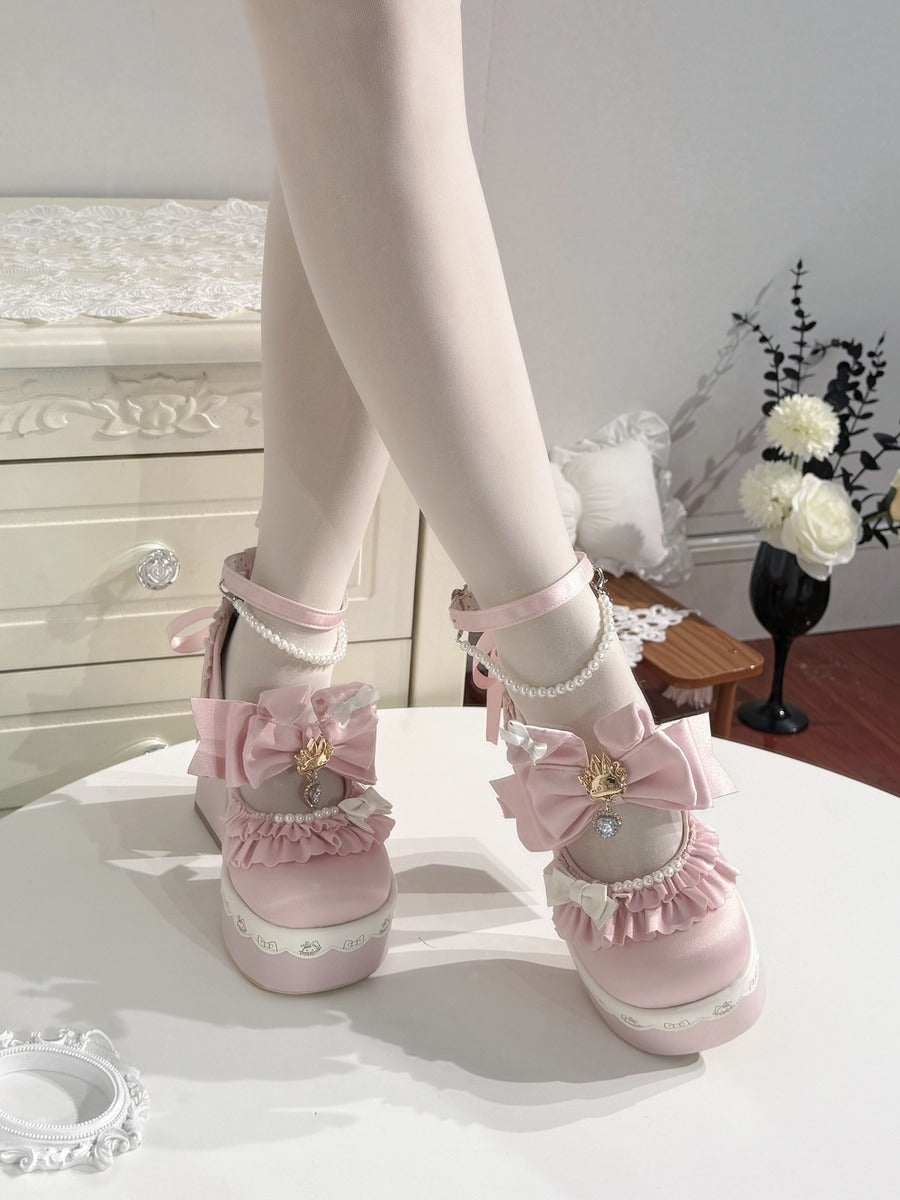 A pair of pastel pink Lolita-style platform shoes with large bows, lace trim, and pearl details, shown on a model's feet standing on a white table in a decorated room.