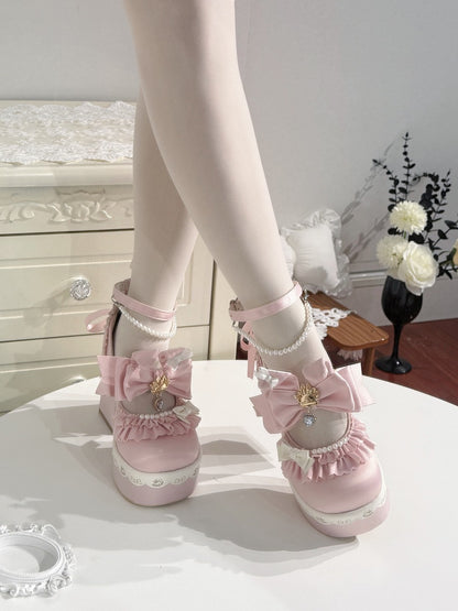 A pair of pastel pink Lolita-style platform shoes with large bows, lace trim, and pearl details, shown on a model's feet standing on a white table in a decorated room.
