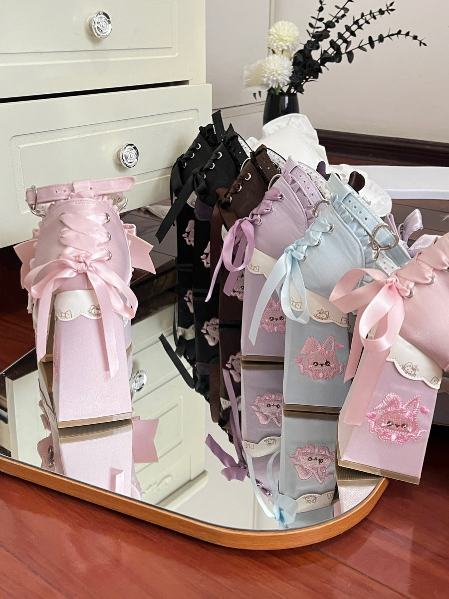A set of pastel-colored lace-up pouches or small bags decorated with large satin ribbons and bows, arranged on a mirrored tray in a room setting with a dresser and flowers in the background.