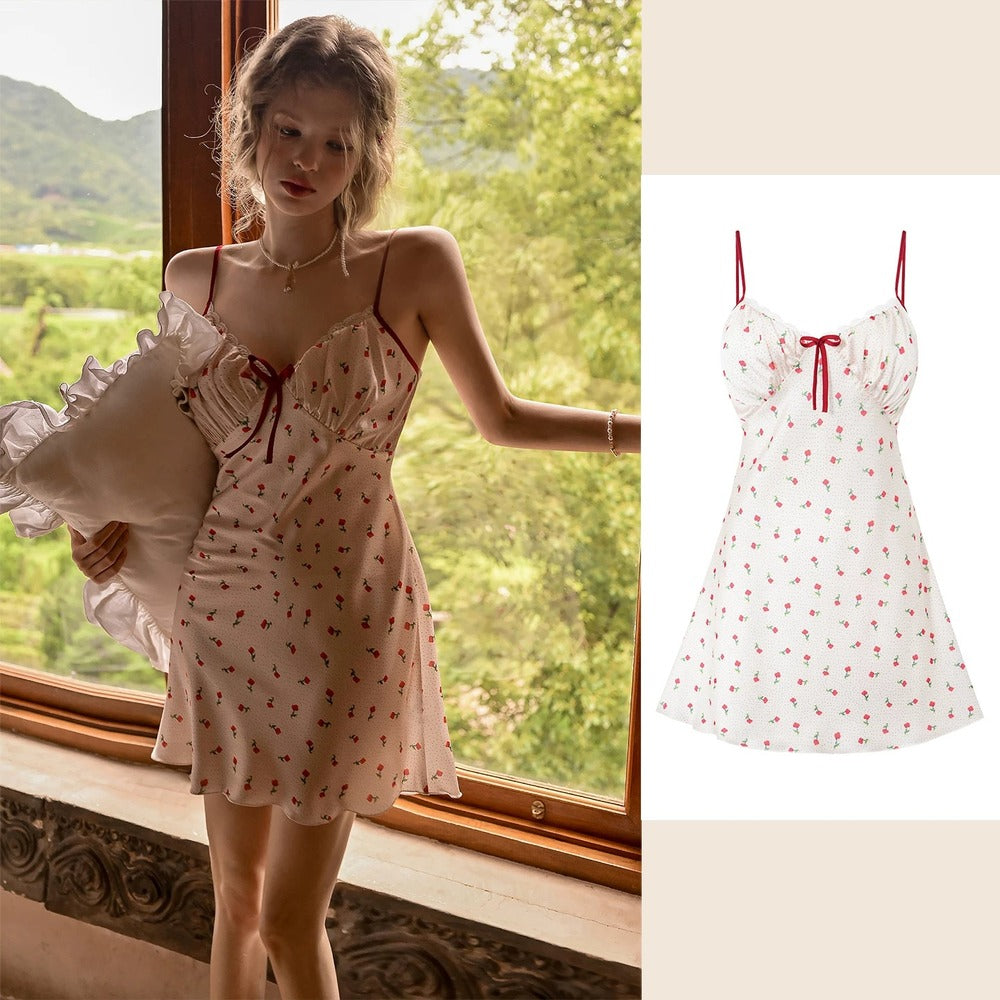 A woman wearing a white spaghetti-strap mini dress with a red cherry print stands by a window holding a pillow, next to a clean product-only view of the dress on the right.