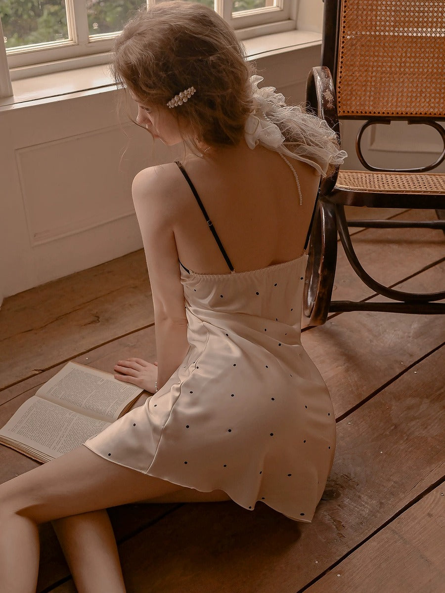 A woman sits on a wooden floor in a warm, vintage-style room, wearing a cream-colored mini dress with small black polka dots and a ruffled white collar. She is seen from behind, looking down at an open book.