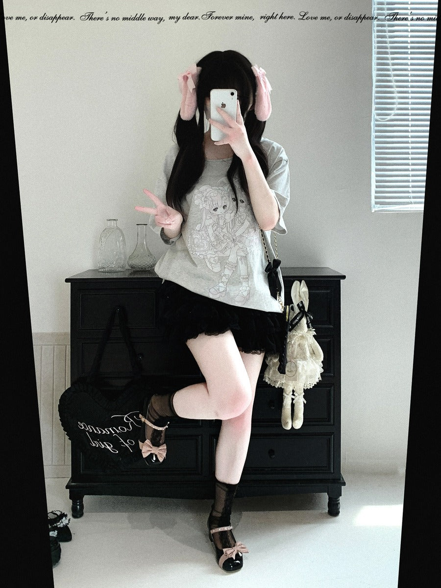 A person takes a mirror selfie in an indoor room, wearing a light gray lace-trimmed short-sleeve top, a black pleated mini skirt, sheer black thigh-high socks, and black strappy heels. A black bag and a white plush toy are placed on a dark wooden cabinet behind them.
