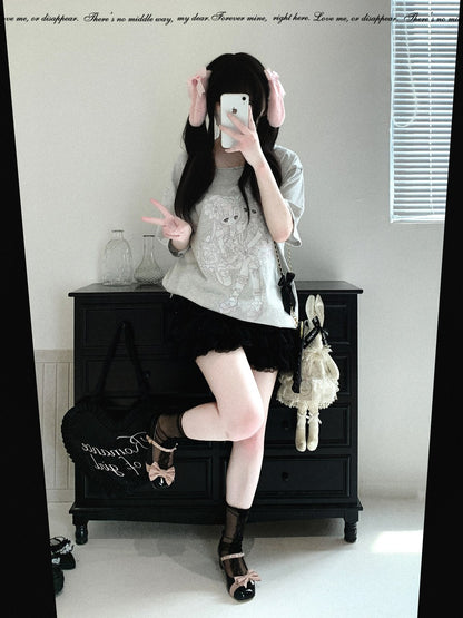 A person takes a mirror selfie in an indoor room, wearing a light gray lace-trimmed short-sleeve top, a black pleated mini skirt, sheer black thigh-high socks, and black strappy heels. A black bag and a white plush toy are placed on a dark wooden cabinet behind them.