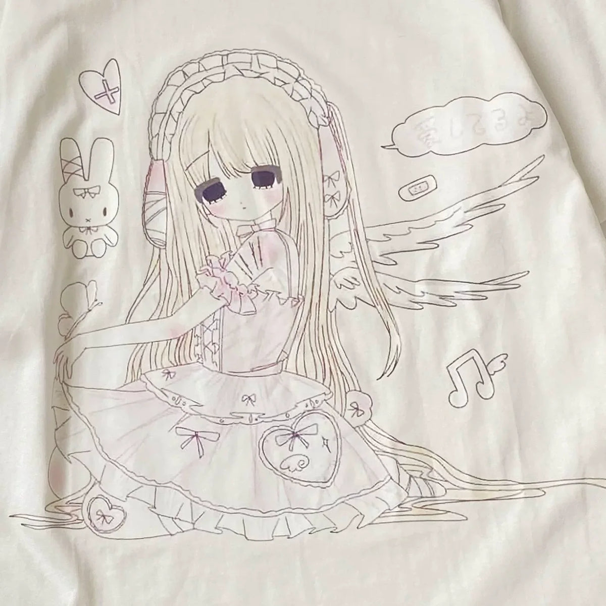 A close-up of a cream-colored garment featuring a detailed anime-style line art graphic of a girl with long blonde hair, a frilly dress, bunny and angel motifs, and Japanese text.