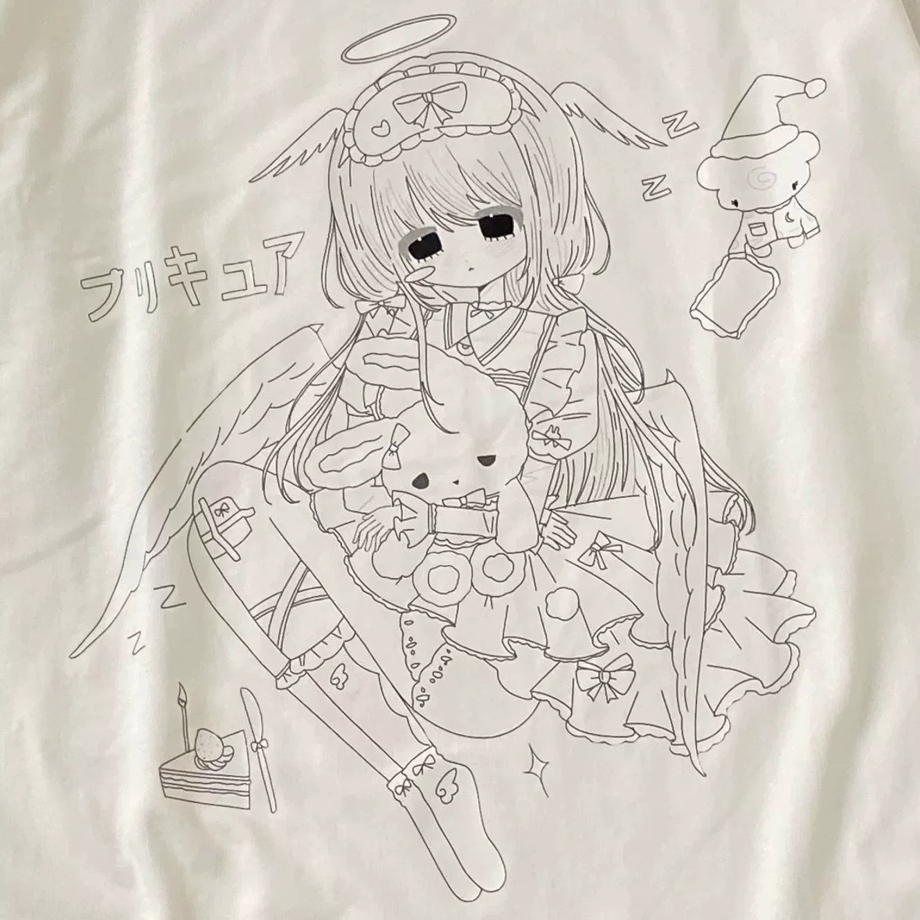 A close-up of a white fabric surface featuring a black line-art graphic of an anime-style angel character holding a rabbit, with small doodle elements and Japanese text.