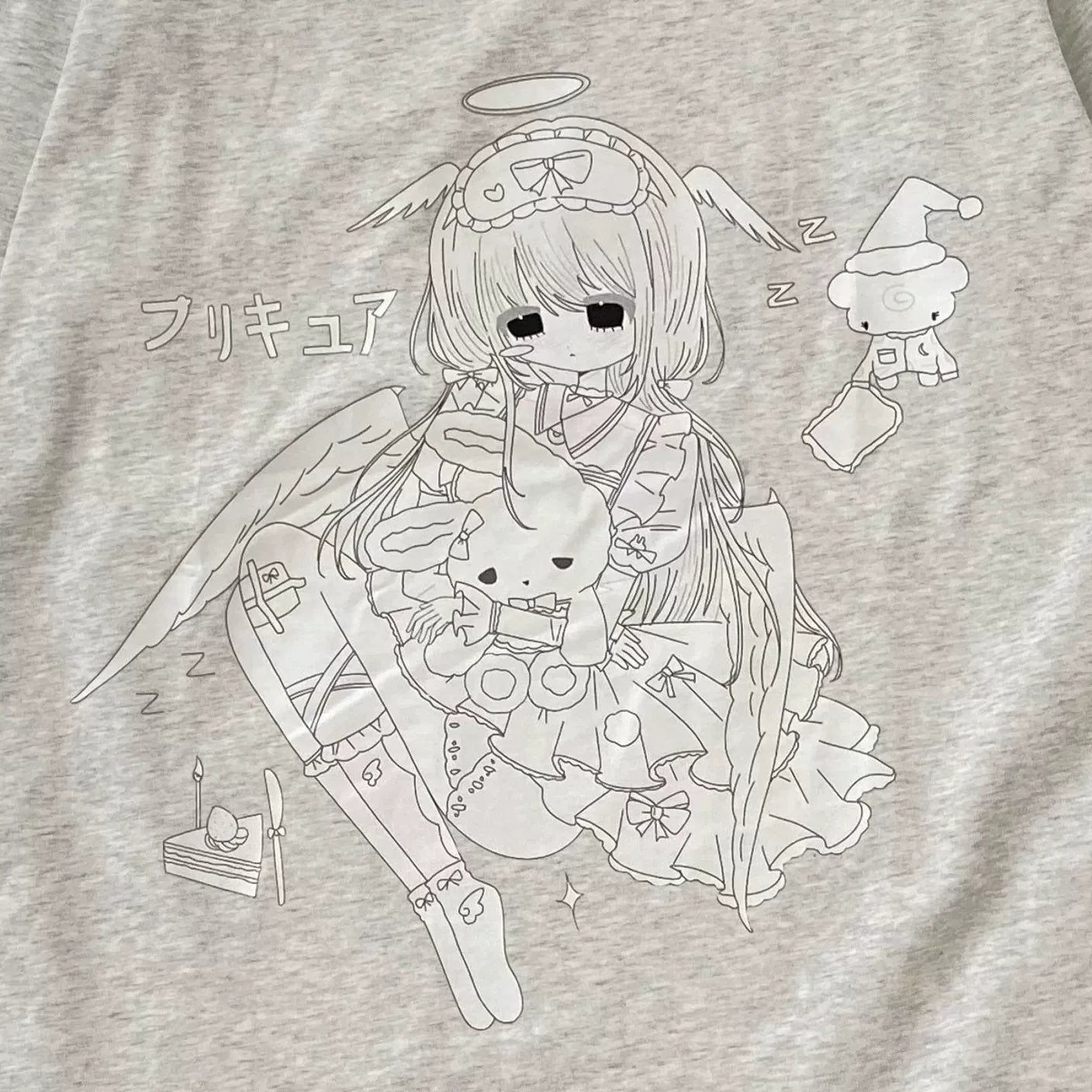Close-up of a light heather gray t-shirt featuring a black line art graphic of a cute anime-style angel girl holding a bunny, with Japanese text and small sleeping character illustrations.