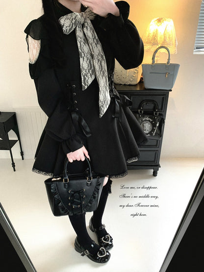 A person wearing a black Lolita-style dress with a large gray and white patterned bow at the collar, holding a black handbag in an indoor room setting.
