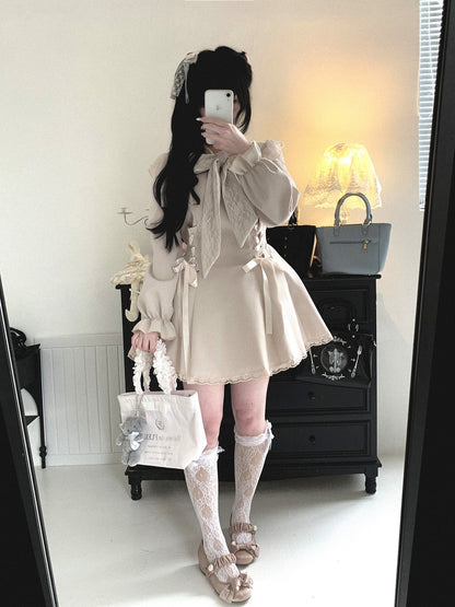 A person takes a mirror selfie in a room wearing a cream-colored Lolita-style dress with lace details, holding a matching cream tote bag. The room has a black wooden chest of drawers, a lit table lamp, and a gray handbag in the background.