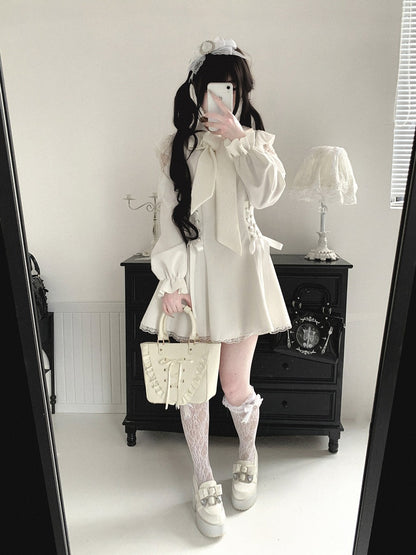 A person takes a mirror selfie in a room, wearing a cream-colored Lolita-style outfit consisting of a long-sleeve blouse with a large bow, a matching mini skirt, lace knee socks, and platform shoes. They are holding a cream-colored quilted handbag.