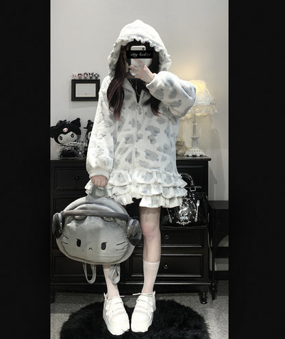 Person wearing a white hooded outfit with ruffles, holding a gray backpack with a cat design, in a room.