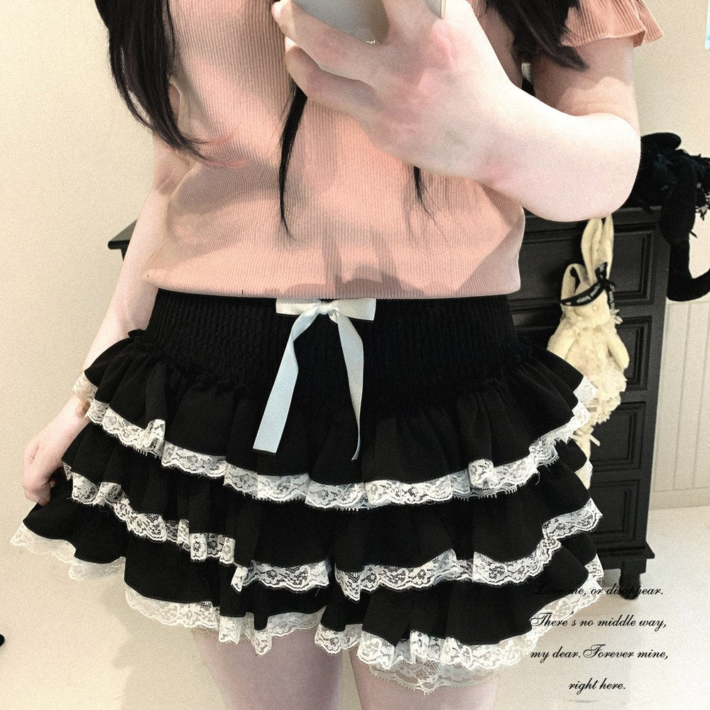 A person wearing a black tiered ruffle skirt with white lace trim and a white bow at the center, paired with a light pink ribbed top, taking a mirror selfie.