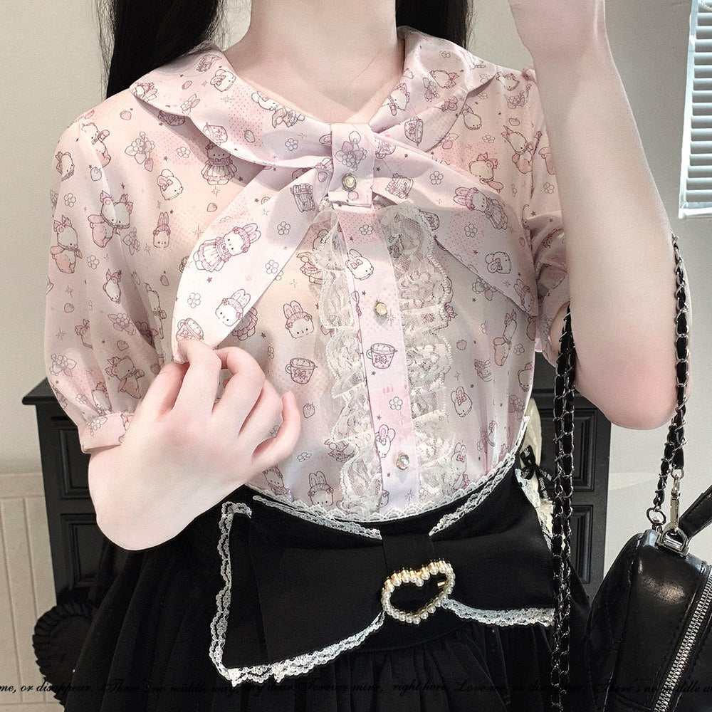 A close-up of a person wearing a light pink short-sleeve blouse with a large bow at the collar, lace trim, and a cartoon print, paired with a black skirt and a black quilted shoulder bag.