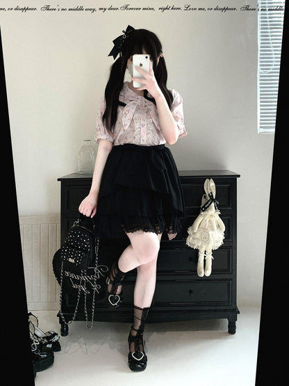 A person taking a mirror selfie in a room, wearing a light pink patterned short-sleeve blouse, a black pleated mini skirt, and black lace-up shoes. They are holding a black studded backpack and standing in front of a black wooden dresser with a cream-colored doll hanging on the side.