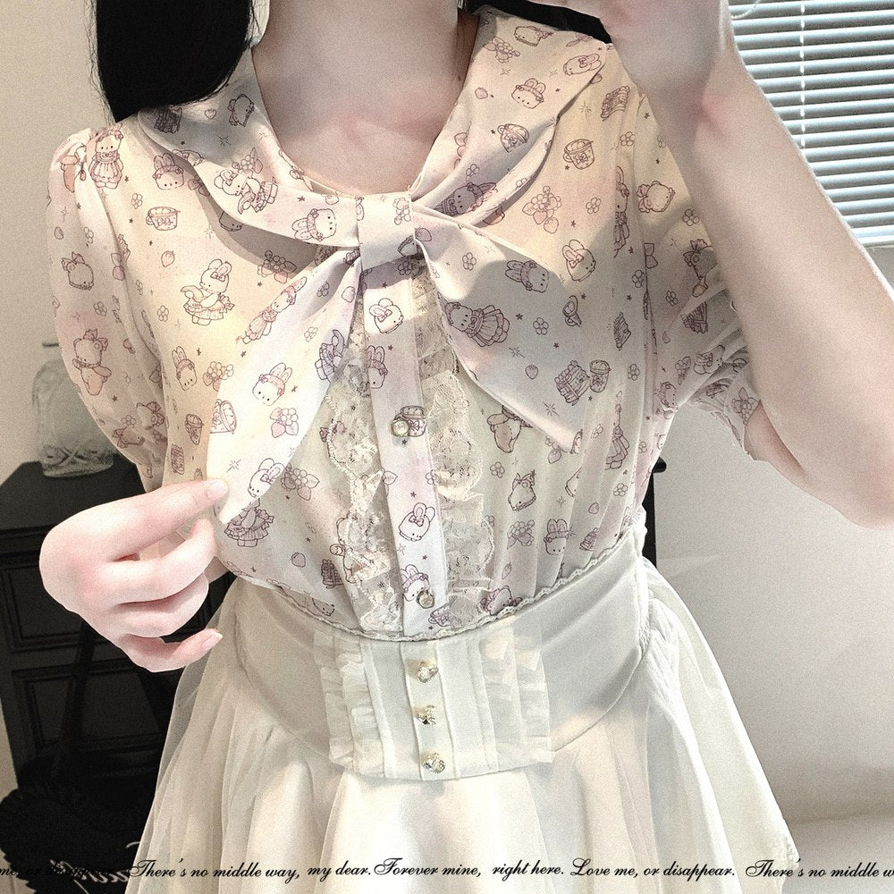 A close-up of a person wearing a short-sleeve blouse with a large bow at the collar, featuring a light beige background with a repeating cute character print in muted pink and brown. The blouse is paired with a cream-colored skirt.