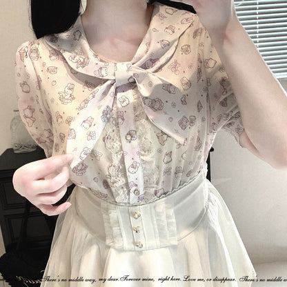 A close-up of a person wearing a short-sleeve blouse with a large bow at the collar, featuring a light beige background with a repeating cute character print in muted pink and brown. The blouse is paired with a cream-colored skirt.