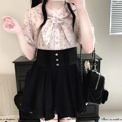 A person wearing a light pink patterned blouse with a large bow at the collar and a black pleated skirt with a wide waistband and front buttons, holding a black quilted shoulder bag in an indoor room.