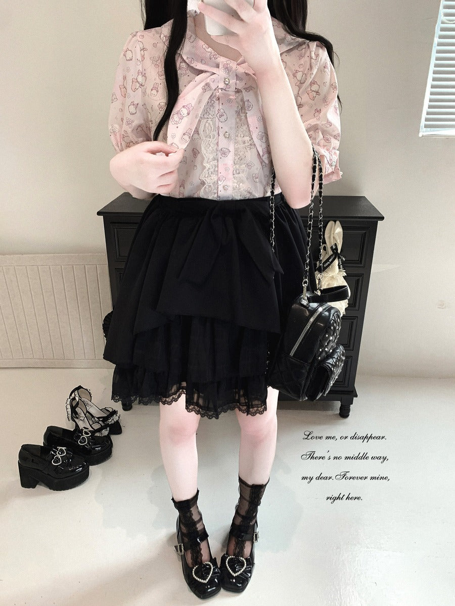 A person wearing a light pink floral short-sleeve blouse with a large bow at the collar, a black tiered ruffled skirt with lace hem, black lace-up Mary Jane shoes, and carrying a small black quilted shoulder bag, standing indoors in front of a dark cabinet.