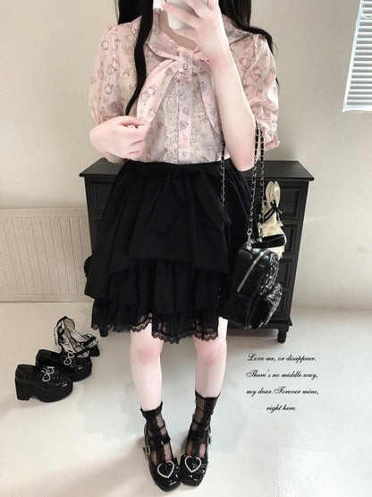 A person wearing a light pink floral short-sleeve blouse with a large bow at the collar, a black tiered ruffled skirt with lace hem, black lace-up Mary Jane shoes, and carrying a small black quilted shoulder bag, standing indoors in front of a dark cabinet.