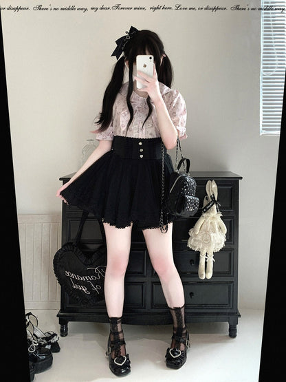 A person takes a mirror selfie wearing a short-sleeve pink lace top, a black pleated mini skirt with silver buttons, sheer black socks, and black lace-up platform shoes. They are carrying a black quilted shoulder bag and holding a black heart-shaped bag in a room with a black wooden chest.