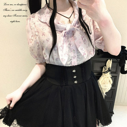 A person wearing a sheer pale pink blouse with a small pastel pattern and a large bow at the neckline, paired with a black high-waisted corset-style skirt. The photo is a mirror selfie in an indoor room with a dark cabinet and hanging bag in the background.