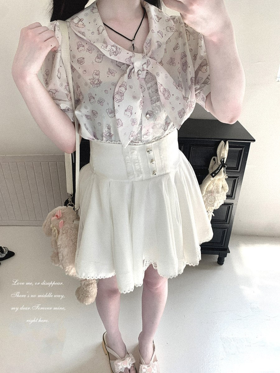 A person wearing a cream-colored short-sleeve blouse with a pink floral print and a matching pleated high-waisted skirt, styled with a small beige plush bag and lace-up shoes in an indoor setting.