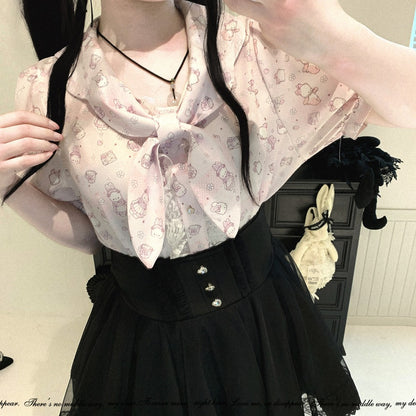 A person wearing a light pink short-sleeve blouse with a small cartoon print and a large bow at the neckline, paired with a black skirt. The photo is taken indoors with a dark cabinet in the background.