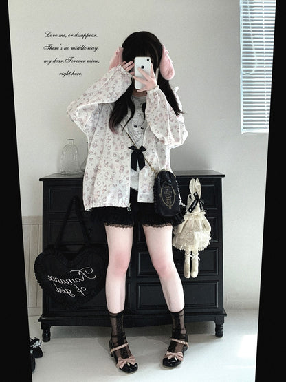 A person takes a mirror selfie showing a fashion outfit consisting of a white floral long-sleeve shirt, black lace shorts, black lace-up sandals, and a black crossbody bag, set in a room with a black wooden chest.