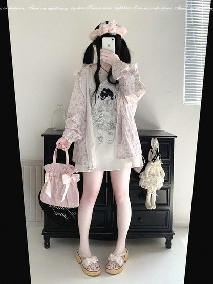 A person takes a mirror selfie in a room, wearing a light pink floral print oversized jacket over a white dress with a black graphic print, holding a matching pink bag with a bow. The setting includes a black wooden dresser in the background.