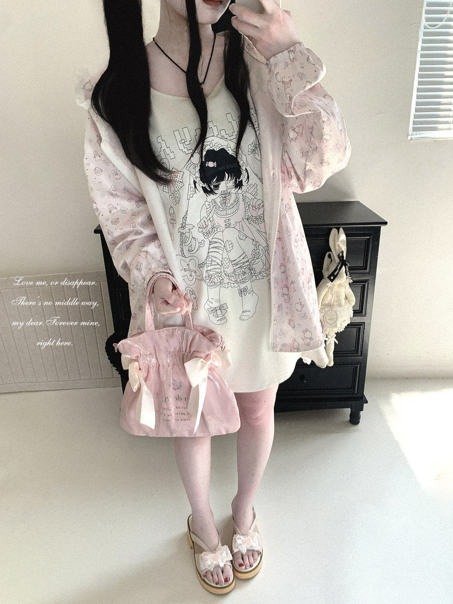 A person modeling a pastel pink floral long-sleeve cardigan over a cream graphic T-shirt, paired with a matching pink handbag and sandals, standing in a room with a black cabinet in the background.