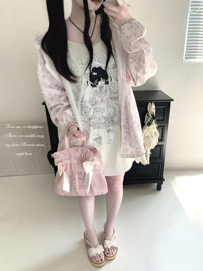 A person modeling a pastel pink floral long-sleeve cardigan over a cream graphic T-shirt, paired with a matching pink handbag and sandals, standing in a room with a black cabinet in the background.