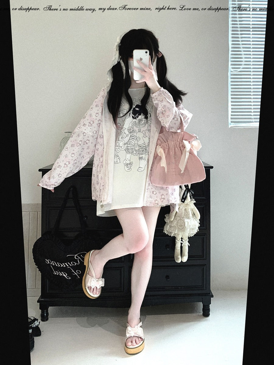 A person takes a mirror selfie in a room, wearing a light pink floral long-sleeve cardigan over a white graphic T-shirt dress, paired with a pink pleated mini skirt, pink shoes, and carrying a pink bag and a small white purse.