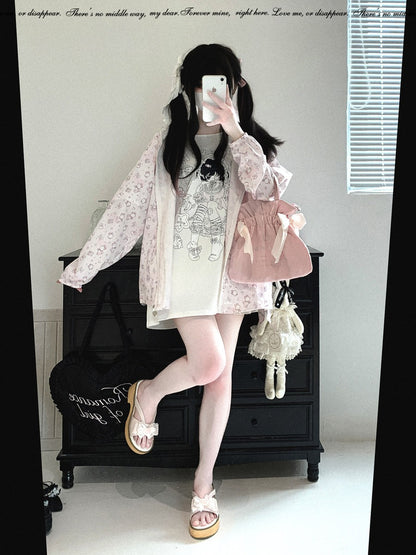 A person takes a mirror selfie in a room, wearing a light pink floral long-sleeve cardigan over a white graphic T-shirt dress, paired with a pink pleated mini skirt, pink shoes, and carrying a pink bag and a small white purse.