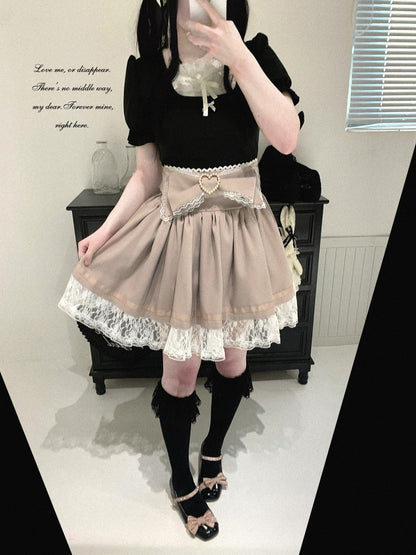 A person wearing a black short-sleeve top and a beige pleated Lolita-style skirt with white lace trim and a heart-shaped belt buckle, paired with black knee-high socks and black shoes with bow details, standing in an indoor room.