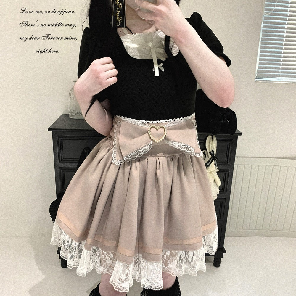 A person wearing a black short-sleeve top and a pleated beige Lolita-style skirt with white lace trim and a heart-shaped belt buckle, standing indoors in front of a dark cabinet.