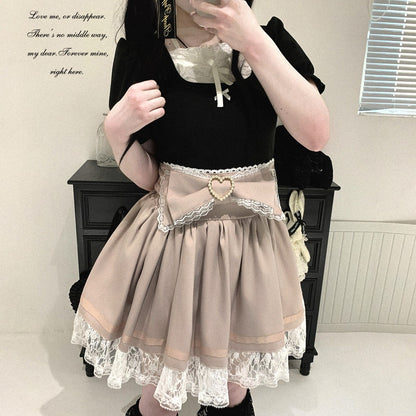 A person wearing a black short-sleeve top and a pleated beige Lolita-style skirt with white lace trim and a heart-shaped belt buckle, standing indoors in front of a dark cabinet.
