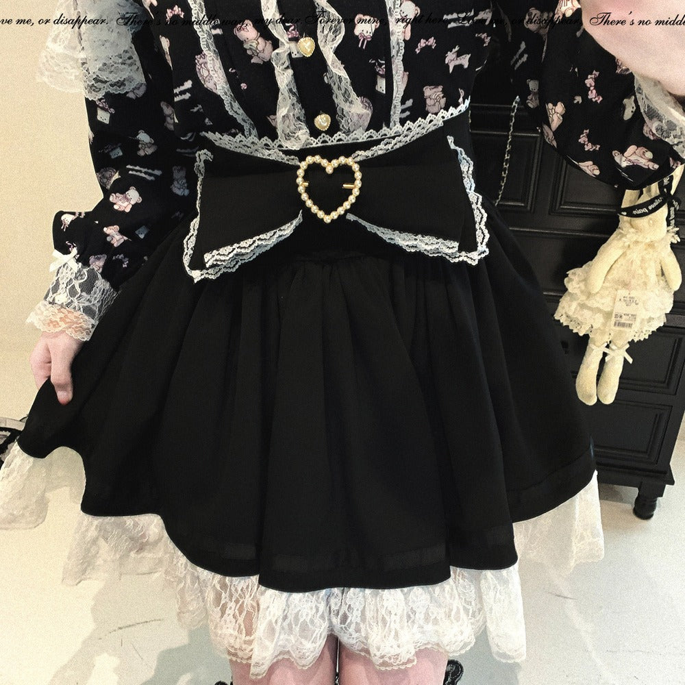 A person wearing a black and white Lolita-style outfit with a black skirt, white lace ruffles, a large black waist bow, and a heart-shaped beaded brooch. The top is black with white lace and small pink floral motifs.