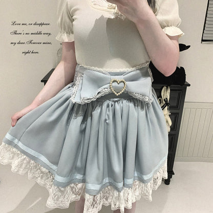 A person wearing a light blue Lolita-style skirt with a large bow, lace trim, and a heart-shaped pearl brooch at the center, paired with a cream top. The photo is taken indoors in front of a dark cabinet.