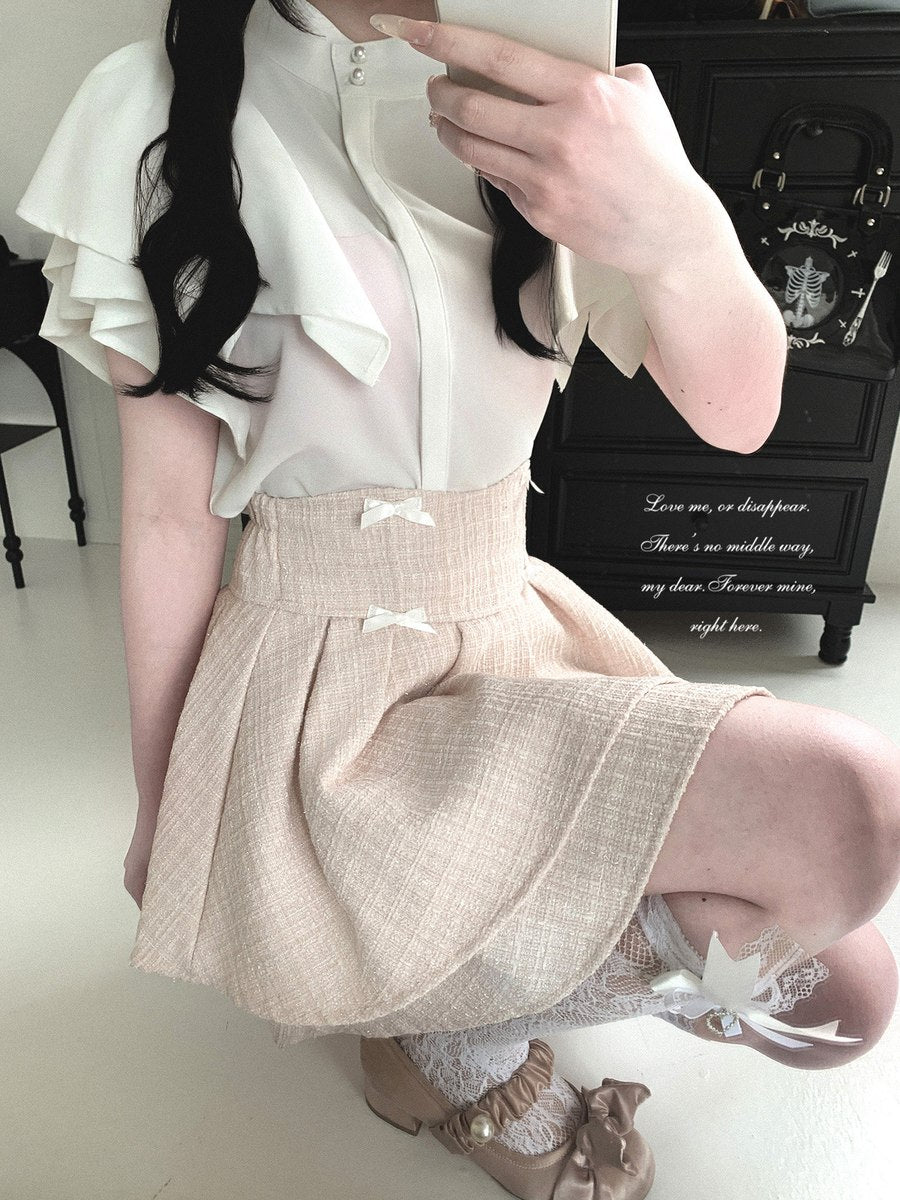 A person wearing a cream ruffled blouse and a pleated beige skirt with small white bows, taking a mirror selfie in an indoor room with a black cabinet in the background.
