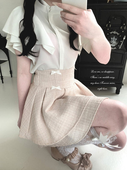 A person wearing a cream ruffled blouse and a pleated beige skirt with small white bows, taking a mirror selfie in an indoor room with a black cabinet in the background.