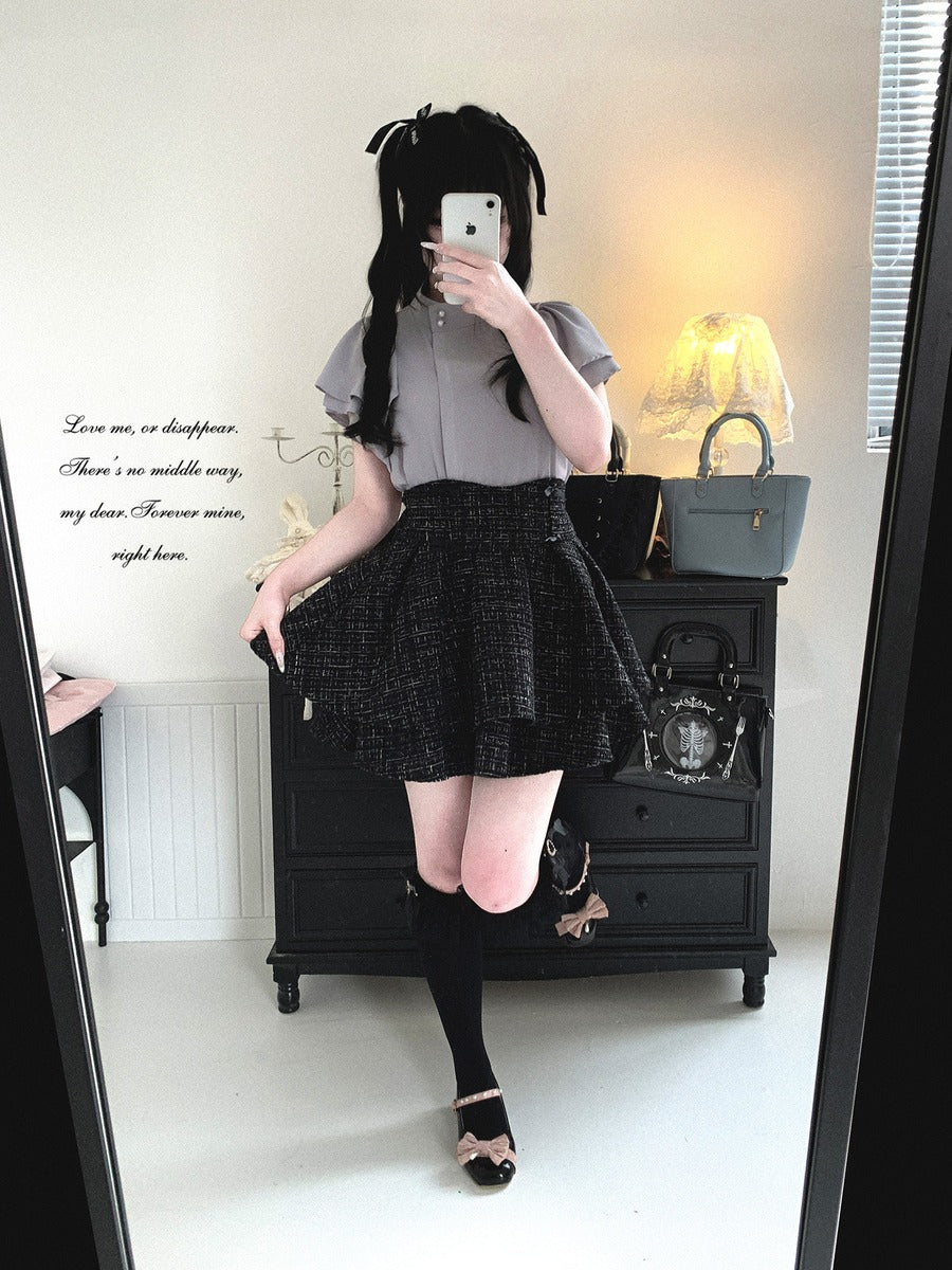 A person taking a mirror selfie in a room, wearing a short-sleeve gray top, a black and white textured pleated mini skirt, black knee-high socks, and black sandals.