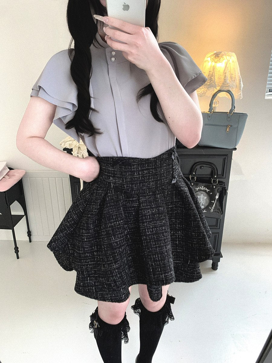 A person taking a mirror selfie wearing a light gray short-sleeve blouse and a black-and-white textured tweed pleated skirt, paired with black knee-high socks.
