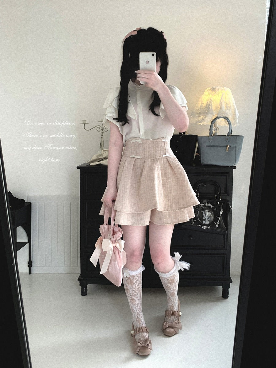 A person taking a mirror selfie in a room, wearing a white short-sleeve blouse, a beige gingham pleated mini skirt, white lace knee-high socks, and beige flat shoes, while holding a pink handbag.