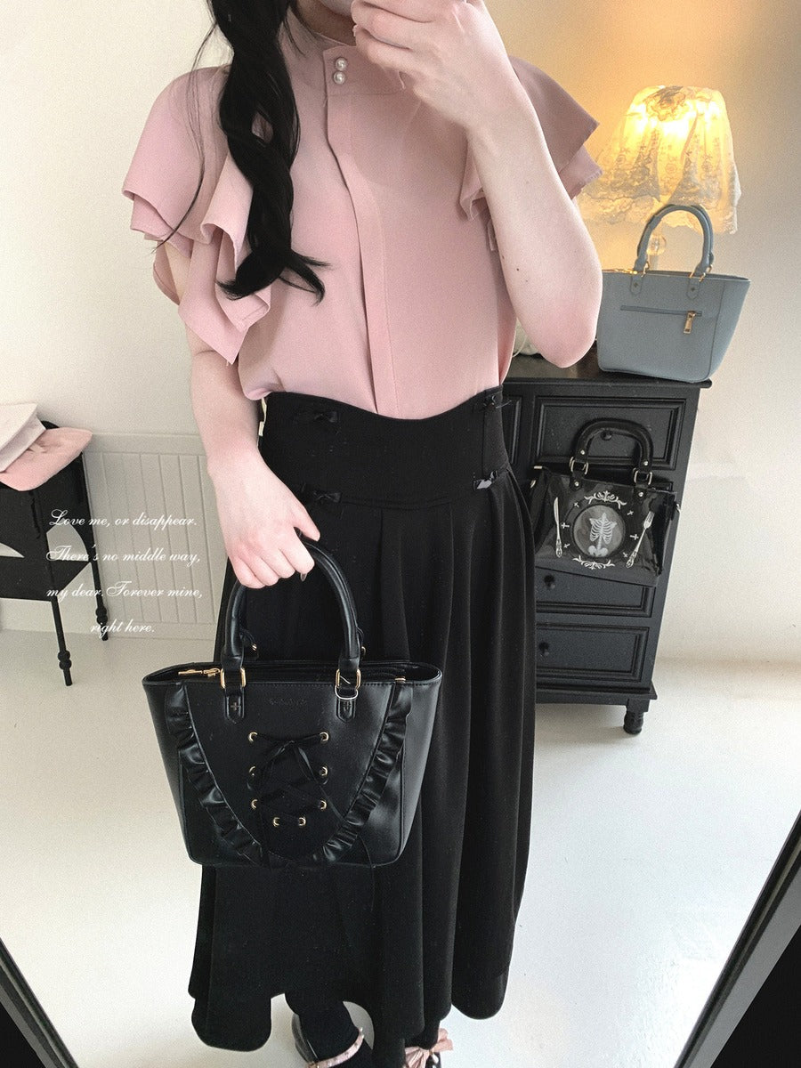 A woman takes a mirror selfie wearing a light pink ruffled short-sleeve blouse and a long black skirt, holding a black lace-up handbag. The background shows a black cabinet with a gray handbag and a lit lamp.