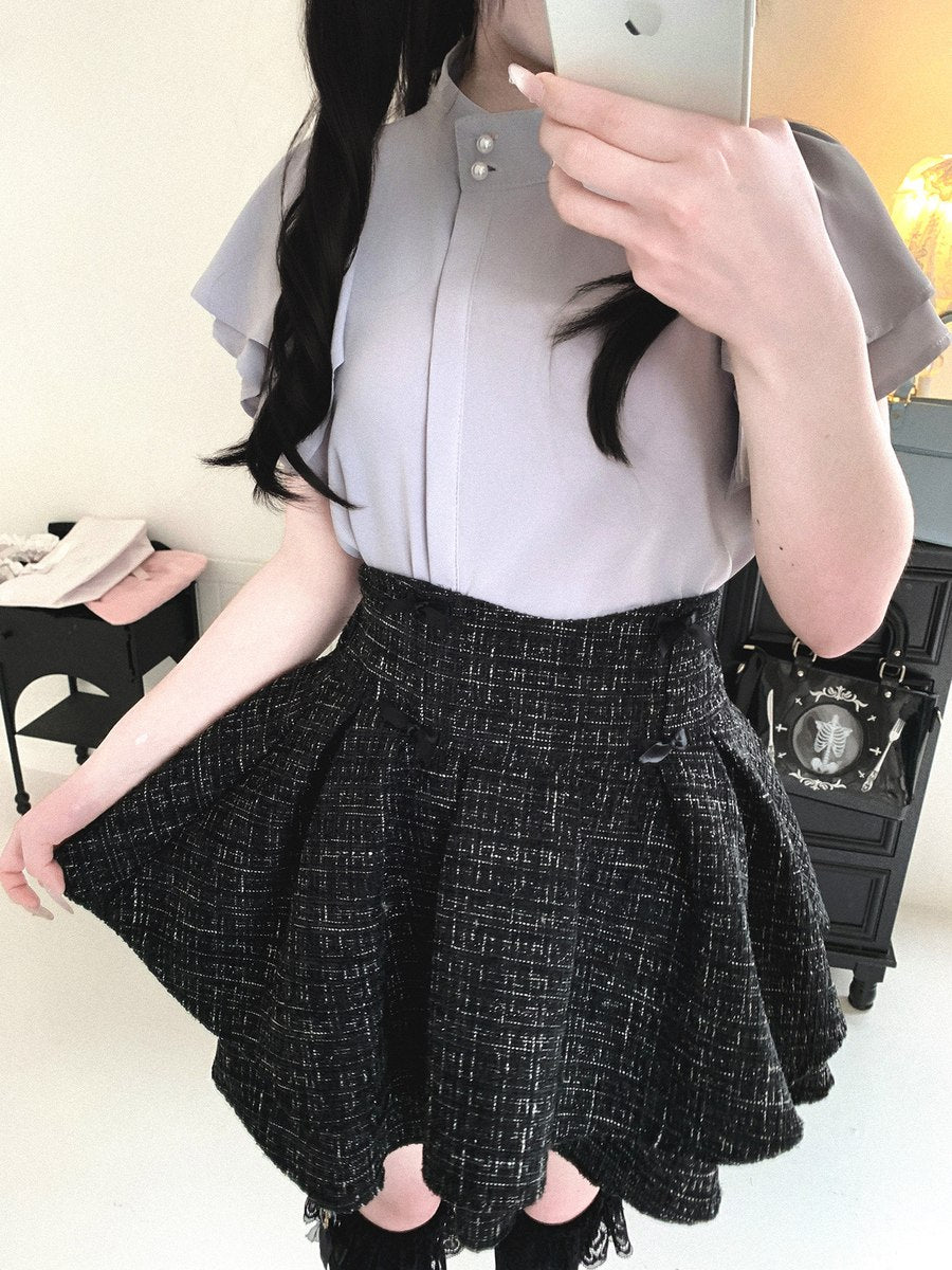 A person taking a mirror selfie wearing a light gray short-sleeve blouse and a black-and-white textured tweed pleated skirt, holding a phone in front of their face.